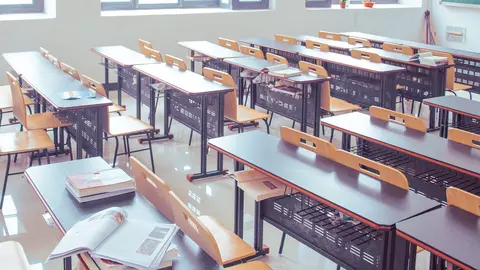 Clase de un colegio con pupitres y sillas y con libros en una de las mesas.